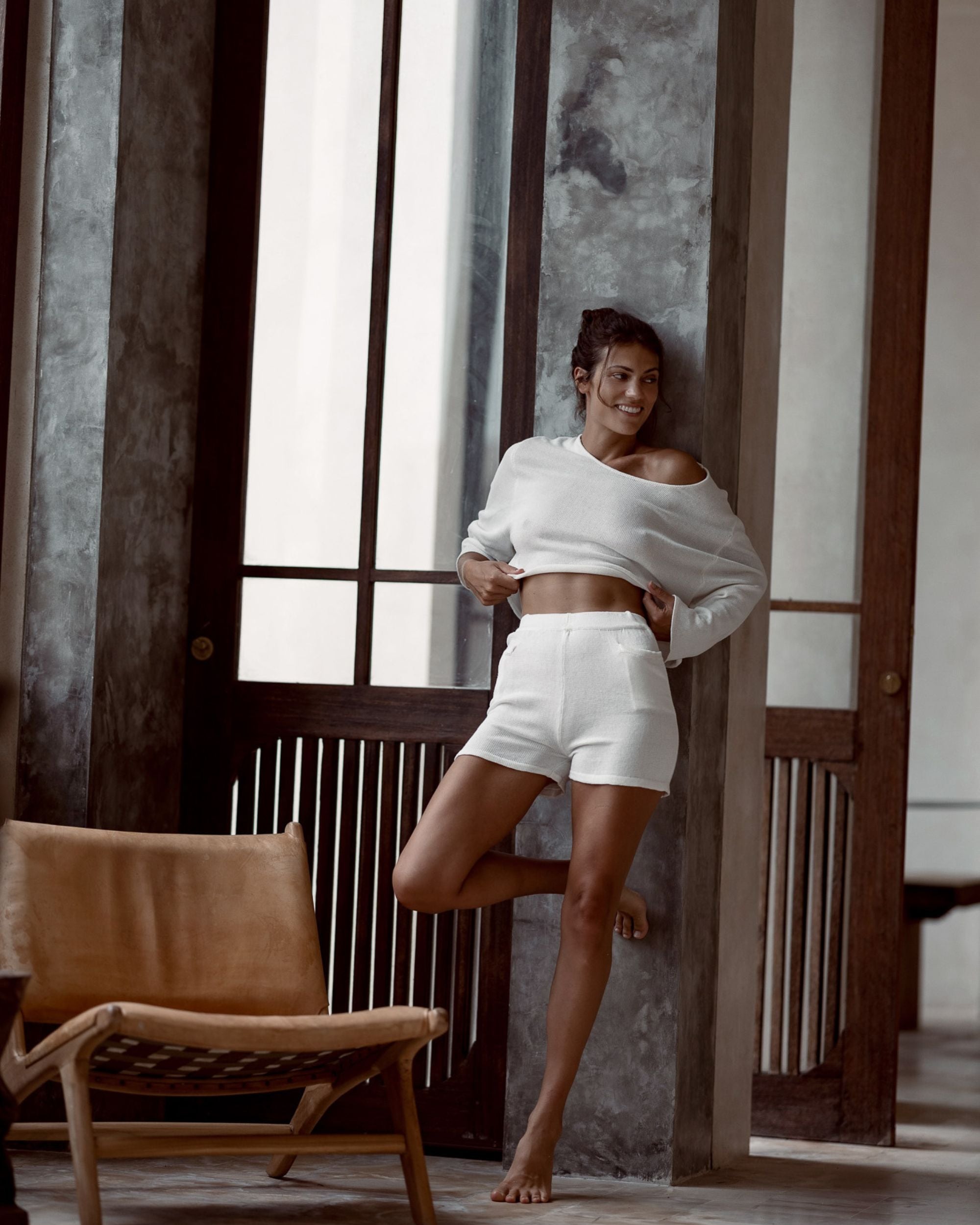Woman in white outfit standing in a room with wooden furniture and large windows.
